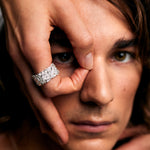 Close-up of a male model with brown hair, making a circular “O” gesture around his eye using his thumb and index finger. He gazes through the loop, drawing attention to the hand gesture. On his index finger is the Ring of Fire Recycled Sterling Silver by Märta Larsson, featuring flame-inspired detailing and a bold 11 mm wide band. The composition emphasizes the ring’s sculptural power and the creative symbolism of the pose.