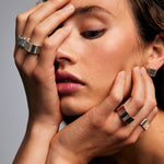 Close-up of a female model’s face against a grey background. One hand rests along the side of her jaw, while the other is placed across the left side of her face. On her middle finger she wears the Ring of Fire Recycled Sterling Silver by Märta Larsson. The ring’s bold 11 mm band and flame-inspired design add a striking, sculptural detail to the intimate and expressive pose