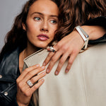 Brown-haired female model embracing another model, both set against a grey background. The focus is on the left hand of the model who is hugging, where she wears the Ring of Fire Recycled Sterling Silver by Märta Larsson on her ring finger. The ring’s flame-inspired detailing and bold 11 mm band add a striking contrast to the soft, intimate moment.