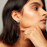 Close-up of a brown-haired female model looking to the right, with one hand resting near her jawline. On her ring finger she wears the MMM Brushed Silver Ring by Märta Larsson. The 10 mm wide band in brushed recycled sterling silver features a continuous row of Märta “M” logos, standing out against the model’s skin and the soft light grey background.