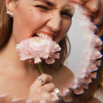 Brown-haired model holding a soft pink flower, playfully appearing to take a bite of it. She wears two Billie Ring Recycled Silver by Märta Larsson—one on the index finger and one on the ring finger. The sculptural recycled silver design contrasts beautifully with the delicate tones, making it a perfect match for silk kaftans and modern caftan fashion.