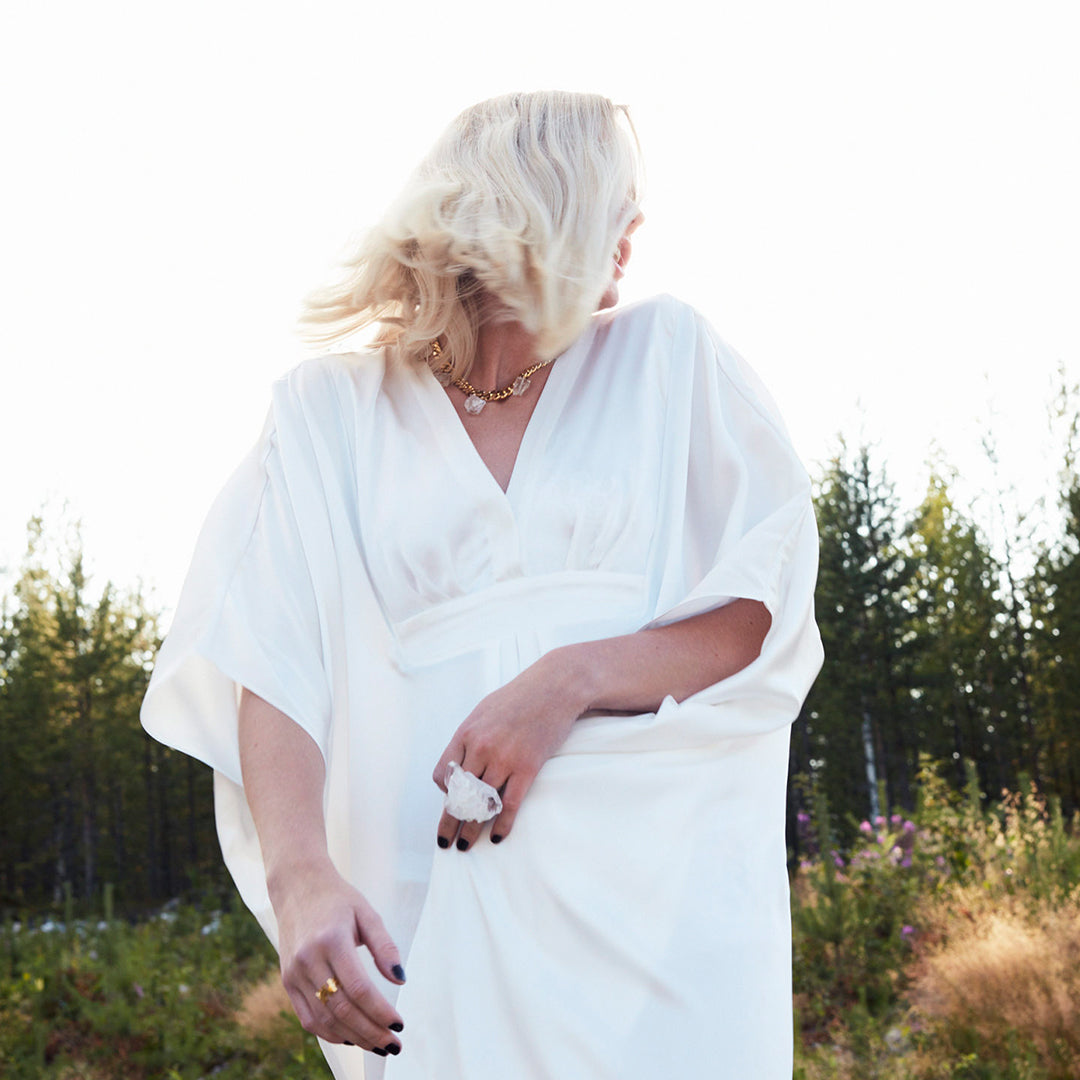 Model in flowing white dress wearing Art Ring Clear Quartz by Märta Larsson. Statement ring styled with gold jewelry in a natural setting — wearable art meets serenity.