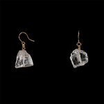 The Raw One Rough Quartz Earrings by Märta Larsson laid out against black background. Clear quartz crystal earrings with gold hooks — bold simplicity and raw brilliance.