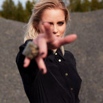Model wearing the Carte Blanche Silk Shirt Dress by Märta Larsson, reaching toward the camera with a forest and gravel background. The dynamic pose and flowing fabric highlight the versatility of this elegant shirt dress for women.