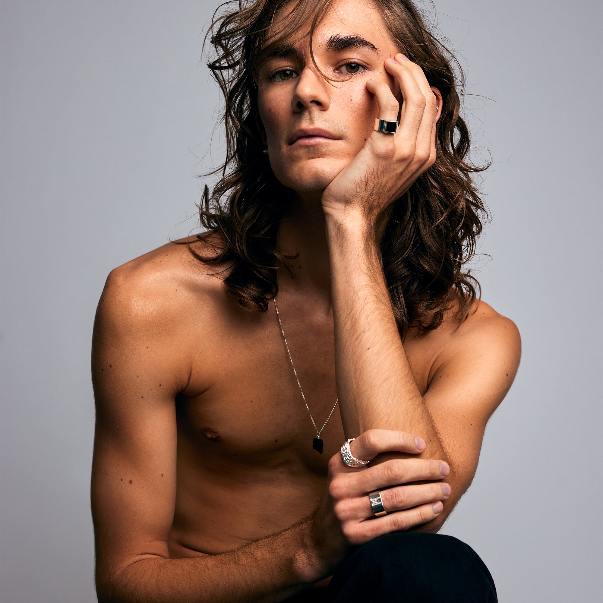 Male model sitting shirtless against a grey background, wearing the Ring of Fire Recycled Sterling Silver by Märta Larsson with bold 11 mm flame-detailed band on his left index finger.