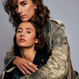 Two models standing against a grey background, the male embracing the female from behind and wearing the Ring of Fire Recycled Sterling Silver by Märta Larsson with bold 11 mm flame-detailed band on his left index finger.