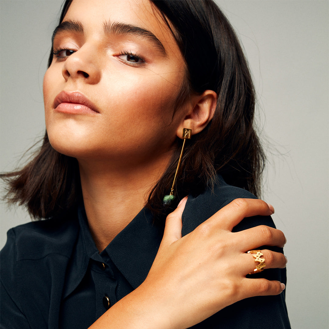  Gold vermeil logo ring worn with dark shirt and green bead earring, showcasing sculptural jewelry styling and confident expression.