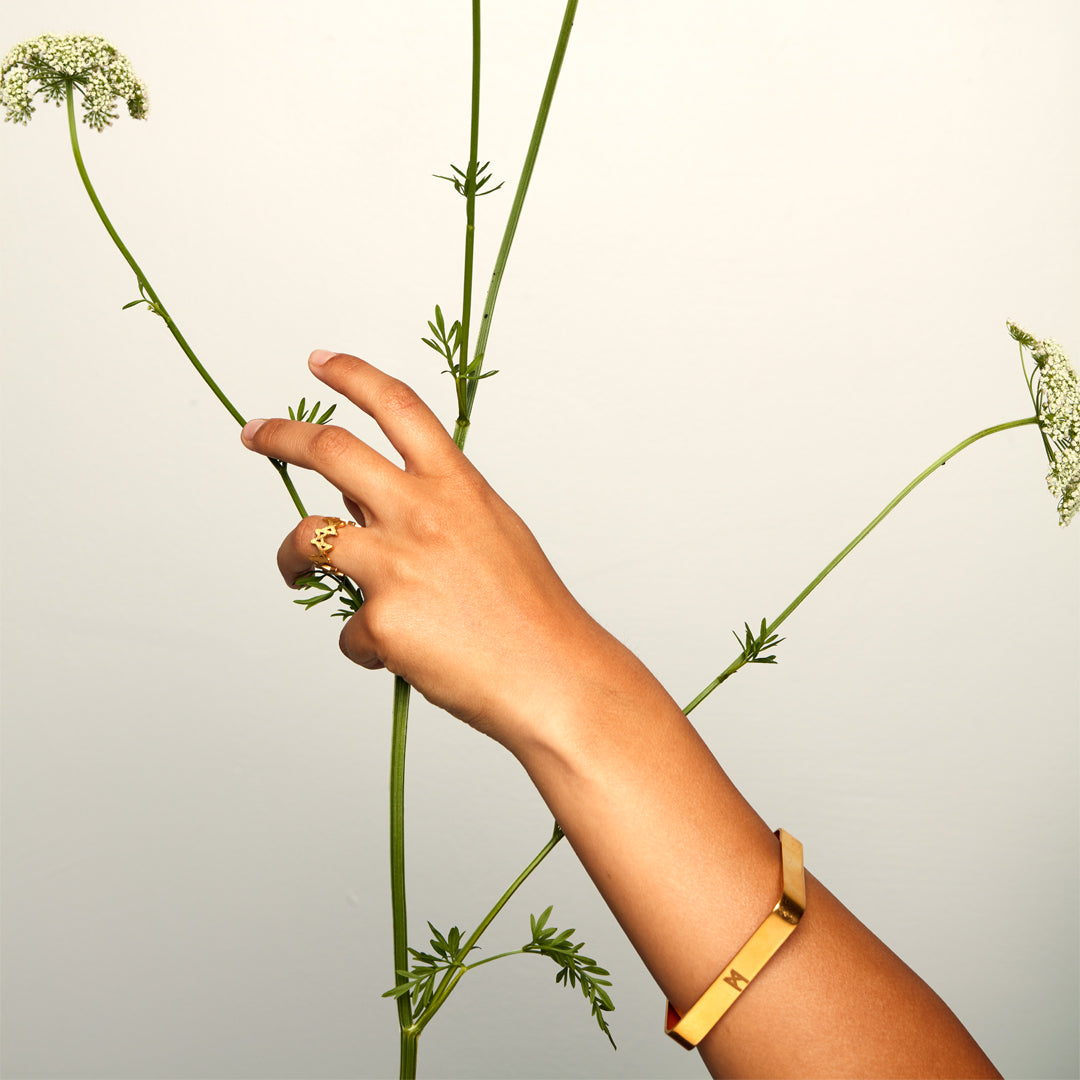 Hand adorned with brushed gold ring and bracelet, gently holding a flowering plant — minimalist styling meets organic elegance.
