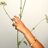 Hand adorned with brushed gold ring and bracelet, gently holding a flowering plant — minimalist styling meets organic elegance.
