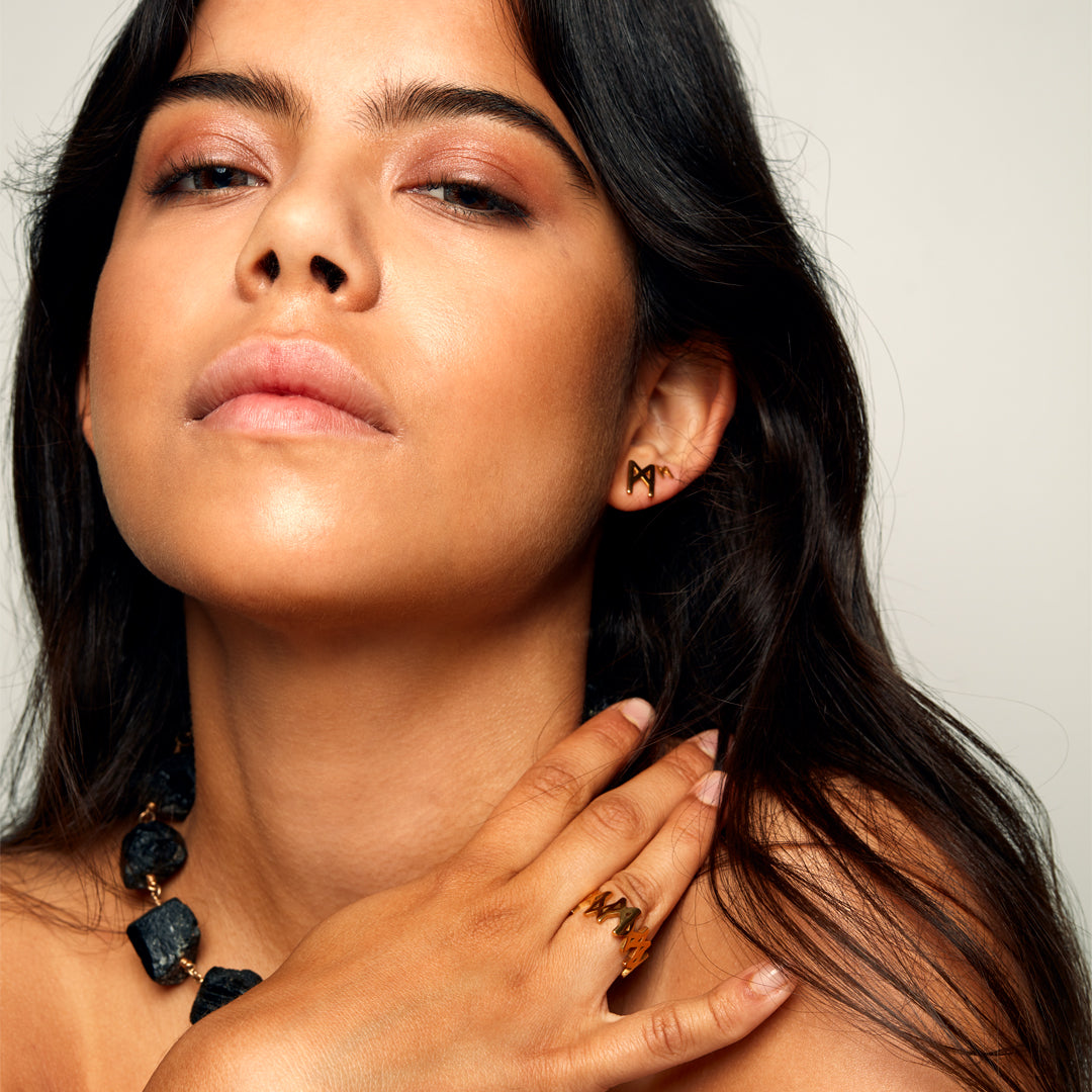 Model with long black hair photographed against a grey background, wearing the MMM Polished Gold Vermeil Ring by Märta Larsson with polished gold surface and repeating “M” logo detail.