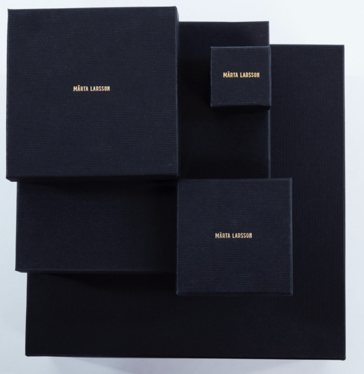 Product packaging for Märta Larsson displayed against a grey background with black boxes of varying sizes and gold lettering, highlighting luxury craftsmanship and refined presentation.