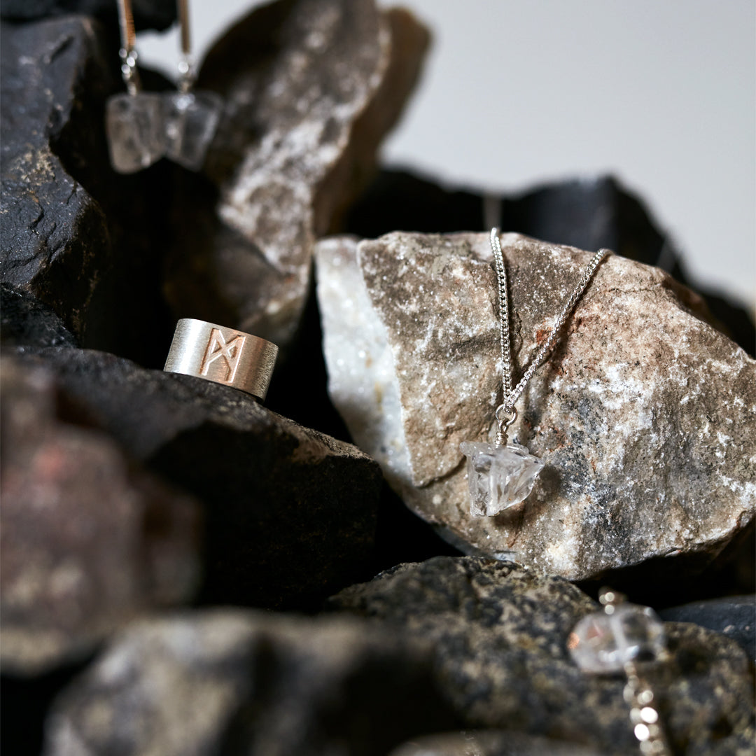 Signature silver ring and crystal necklaces displayed on raw stones, showcasing contrast between rugged texture and refined craftsmanship.