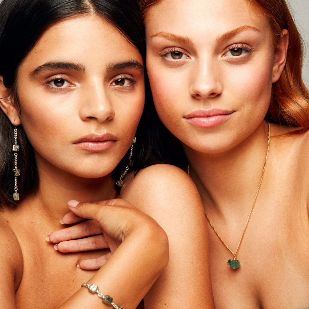 Two models standing side by side, with the ginger-haired model on the right wearing The Raw One Necklace by Märta Larsson featuring a gold chain and raw green fluorite stone.