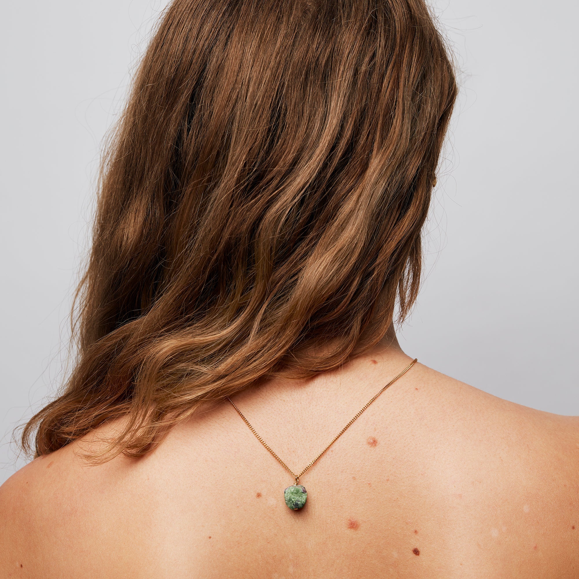 Brown hair woman with her hair swept to one side, showcasing The Raw One Necklace by Märta Larsson with the gold chain and ruby zoisite stone hanging down her spine.