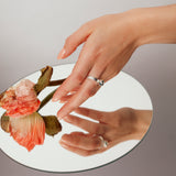 Hand reaching over a mirror to pick up an apricot-colored flower, wearing the Billie Ring Recycled Silver by Märta Larsson with raw cast stone and organic refined texture.