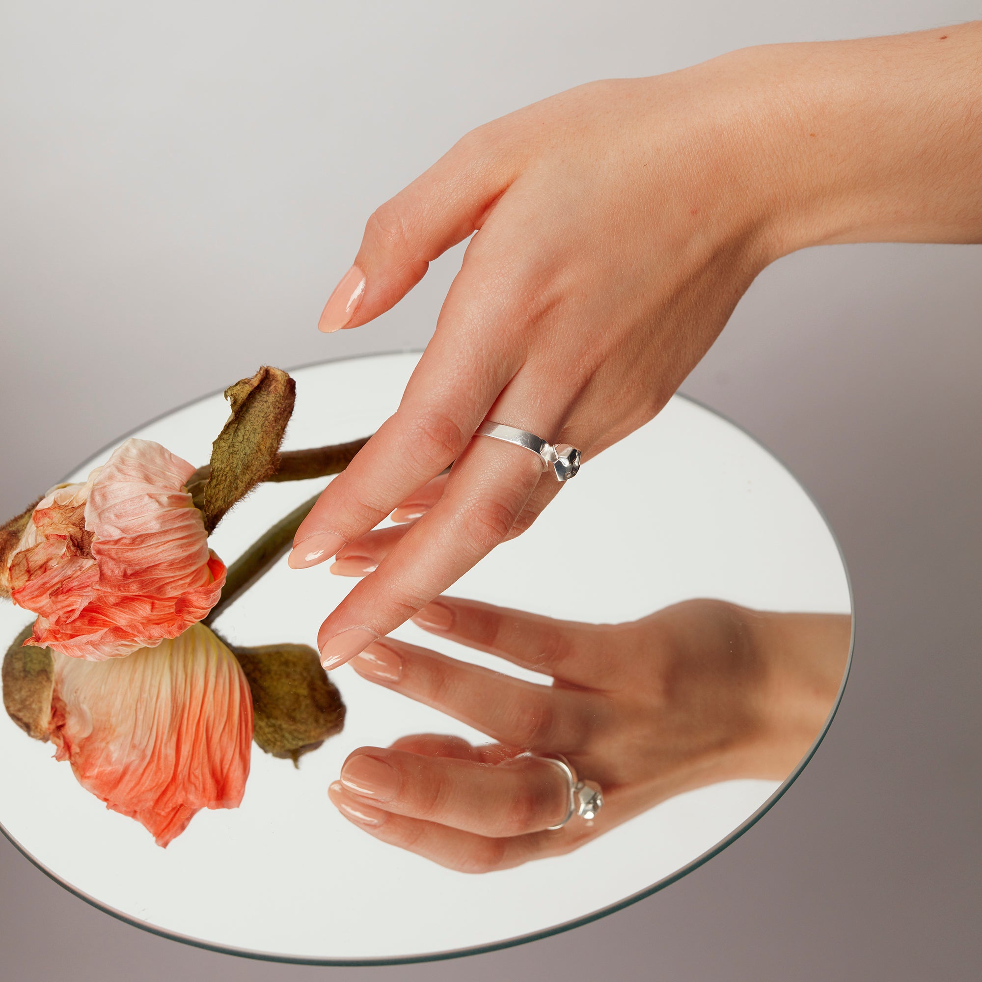 Hand reaching over a mirror to pick up an apricot-colored flower, wearing the Billie Ring Recycled Silver by Märta Larsson with raw cast stone and organic refined texture.