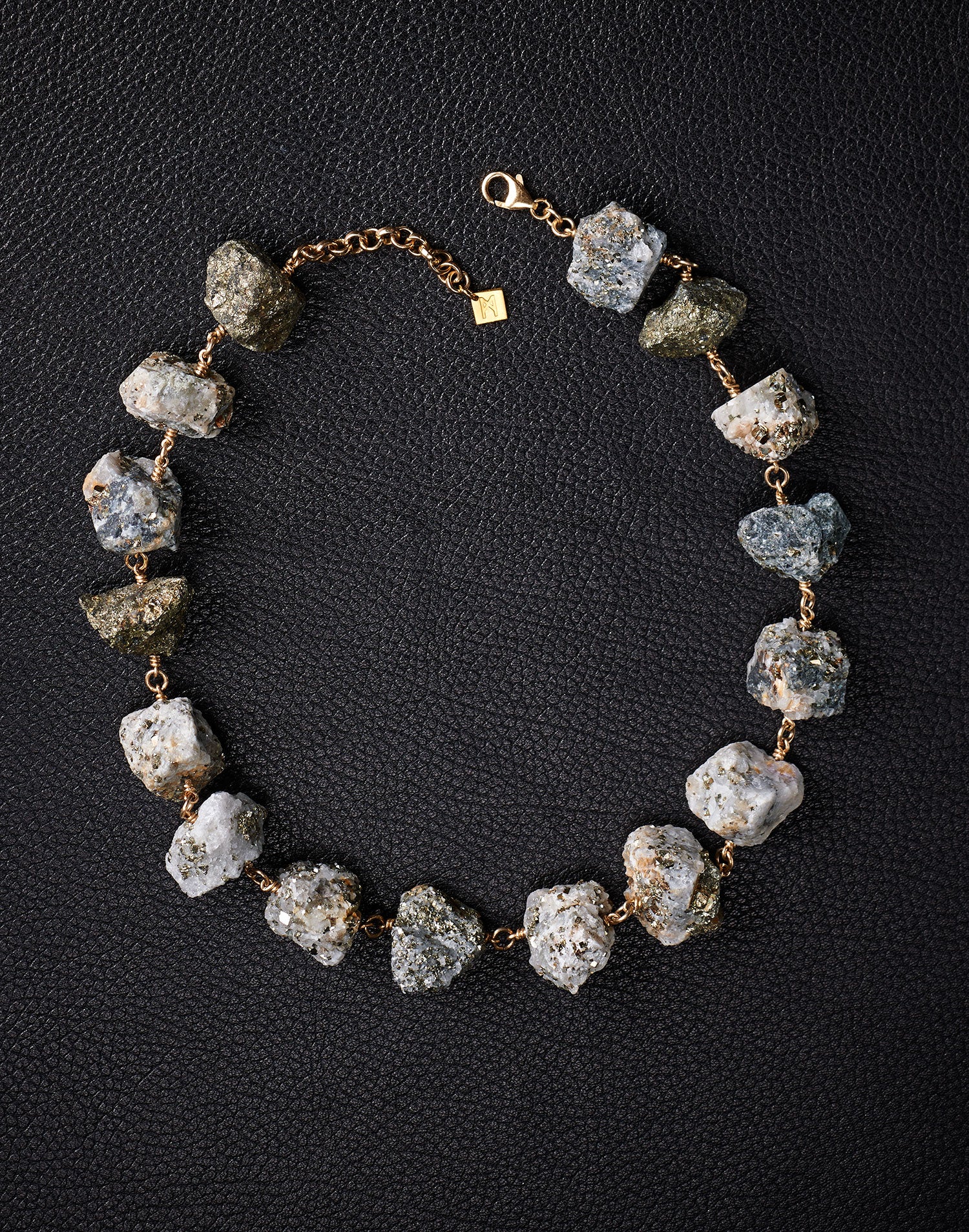 Not a Pearl Necklace Calcite Pyrite by Märta Larsson laid out on a black textured surface, featuring raw calcite-pyrite stones on a gold chain.