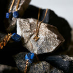Signature Necklace Polished Gold Vermeil by Märta Larsson displayed on textured rocks, an M-logo charm necklace styled with lapis lazuli and gold accents.