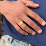 Zoomed-in image of a male hand wearing the Signature Brushed Gold Ring by Märta Larsson in brushed 24 carat gold vermeil with wide matte band against textured dark blue fabric.